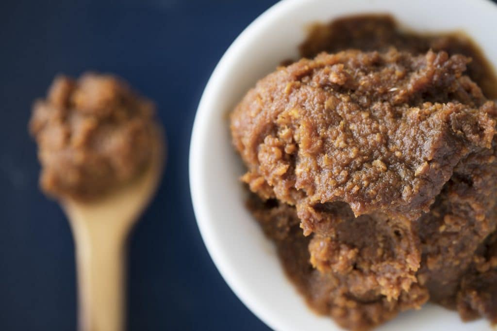 Red miso paste on white bowl viewed from above with spoon on table out of focus.