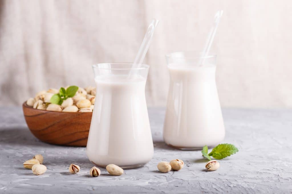 Organic non dairy pistachio milk in glass and wooden plate with pistachio nuts on a gray concrete background. Vegan healthy food concept, close up, side view.